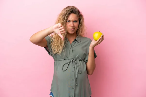 Girl with curly hair isolated on pink background pregnant and holding an apple while doing bad signal