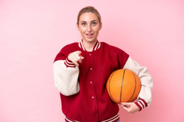 Genç basketbolcu beyaz kadın pembe arka planda izole edilmiş şaşırmış ve ön tarafı işaret ediyor.