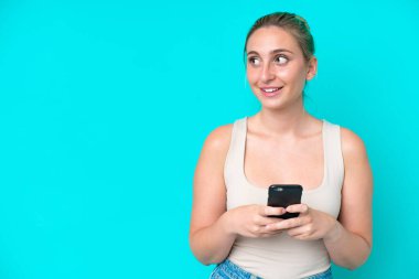 Kafkasyalı genç bir kadın mavi arka planda izole edilmiş ve cep telefonu kullanıyor.