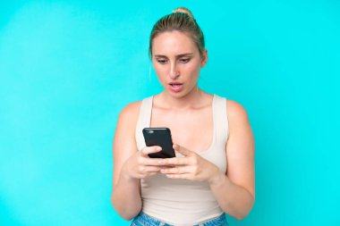 Mavi arka planda izole edilmiş genç beyaz kadın kameraya bakarken şaşırmış bir ifadeyle cep telefonunu kullanıyor.