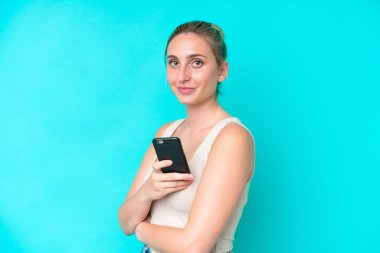 Mavi arka planda izole edilmiş genç beyaz bir kadın cep telefonu tutuyor ve kollarını kavuşturuyor.