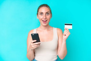Kafkasyalı genç bir kadın mavi arka planda izole edilmiş cep telefonuyla alışveriş yapıyor ve elinde şaşırmış bir ifadeyle bir kredi kartı tutuyor.