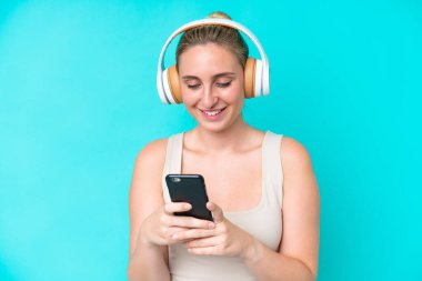 Genç beyaz kadın mavi arka planda izole edilmiş müzik dinliyor ve cep telefonu arıyor.