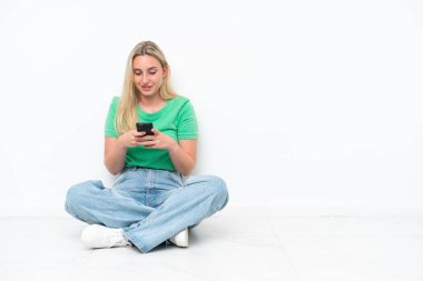 Beyaz arka planda izole bir şekilde yerde oturan beyaz bir kadın cep telefonuyla bir mesaj gönderiyor.