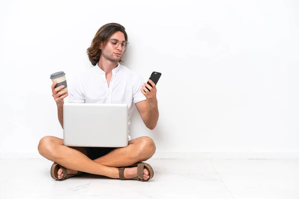 Young handsome man with a laptop sitting on the floor isolated on white background holding coffee to take away and a mobile