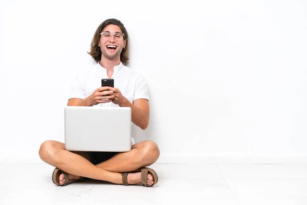 Young handsome man with a laptop sitting on the floor isolated on white background surprised and sending a message