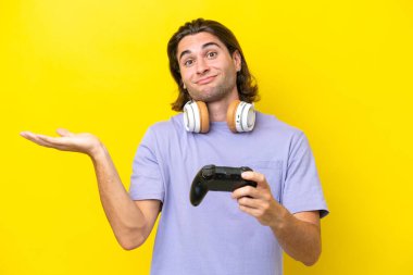 Young handsome caucasian man playing with a video game controller over isolated on yellow background having doubts while raising hands