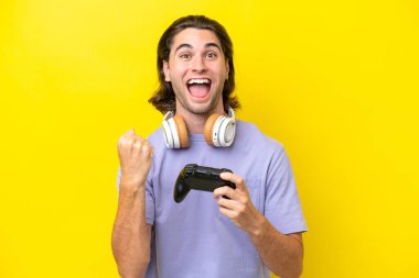 Young handsome caucasian man playing with a video game controller over isolated on yellow background celebrating a victory in winner position