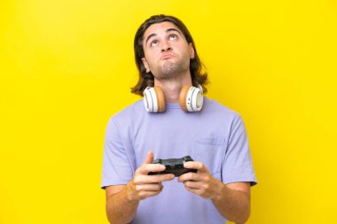 Young handsome caucasian man playing with a video game controller over isolated on yellow background and looking up
