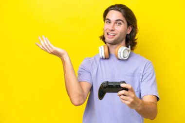 Young handsome caucasian man playing with a video game controller over isolated on yellow background extending hands to the side for inviting to come