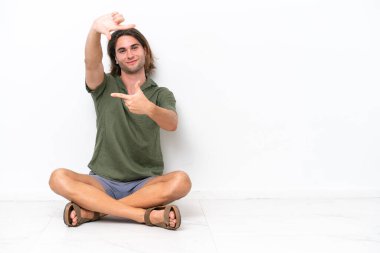 Young handsome man sitting on the floor isolated on white background focusing face. Framing symbol