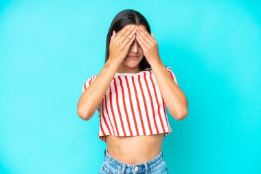 Mavi arka planda izole edilmiş genç beyaz kadın gözlerini elleriyle kapatıyor.