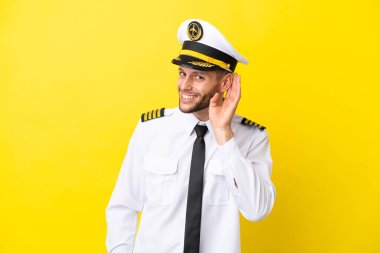 Kafkasyalı uçak pilotu sarı arka planda izole edilmiş bir şekilde elini kulağına koyarak bir şey dinliyor.