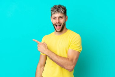 Mavi arka planda izole edilmiş yakışıklı beyaz adam şaşırmış ve işaret ediyor.