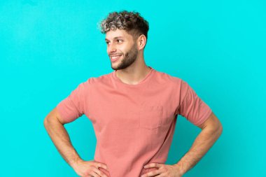 Mavi arka planda soyutlanmış yakışıklı beyaz adam kolları kalçasında ve gülümsüyor.