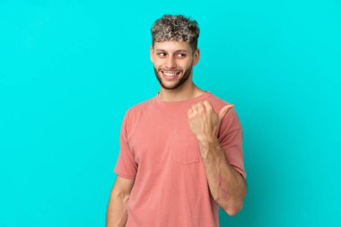 Mavi arka planda izole edilmiş yakışıklı beyaz adam bir ürünü sunmak için kenarı işaret ediyor.