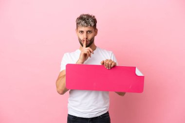 Young handsome caucasian man isolated on pink background holding an empty placard doing silence gesture