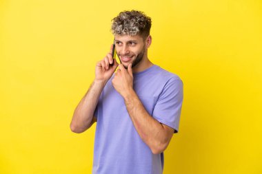 Sarı arka planda izole edilmiş cep telefonu kullanan genç beyaz adam yana bakıyor ve gülümsüyor.