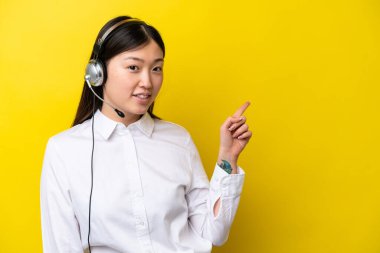 Telemarketer Chinese woman working with a headset isolated on yellow background pointing finger to the side