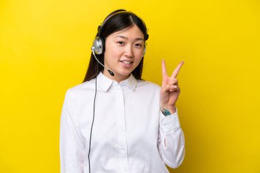 Telemarketer Chinese woman working with a headset isolated on yellow background smiling and showing victory sign