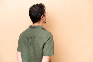 Young caucasian man isolated on beige background in back position and looking back