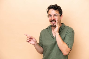 Young caucasian man isolated on beige background pointing to the side to present a product and whispering something