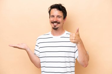 Young caucasian man isolated on beige background holding copyspace imaginary on the palm to insert an ad and with thumbs up