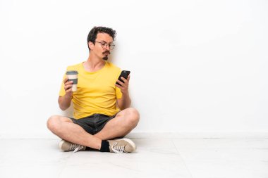 Beyaz arka planda izole bir şekilde yerde oturan genç beyaz bir adam elinde kahve ve bir cep telefonu tutuyor.