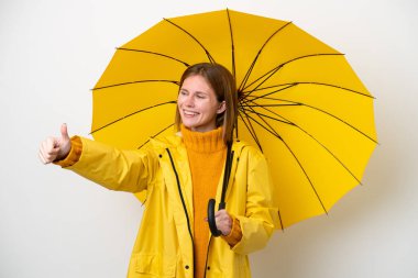 Yağmura dayanıklı ceketi ve şemsiyesi olan genç bir İngiliz kadın beyaz arka planda izole edilmiş bir şekilde baş parmak işareti yapıyor.