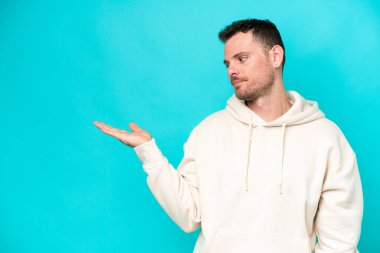 Mavi arka planda izole edilmiş beyaz, yakışıklı bir genç. Kopyalama alanını şüphe ile tutuyor.