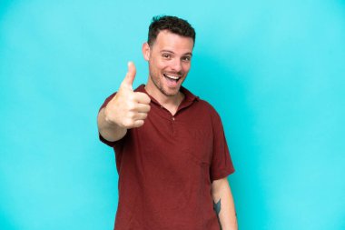 Beyaz tenli yakışıklı adam mavi arka planda izole edildi. Başparmakları havada, çünkü iyi bir şey oldu.