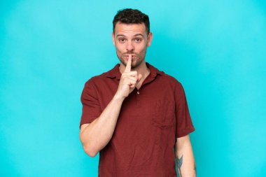 Mavi arka planda izole edilmiş beyaz, yakışıklı genç bir adam sessiz bir jest yapıyor. Parmağını ağzına sokuyor.