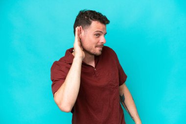 Beyaz tenli yakışıklı adam mavi arka planda izole edilmiş bir şekilde elini kulağına koyarak bir şeyler dinliyor.