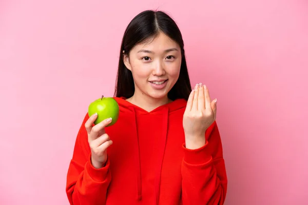 Young Chinese woman with an apple isolated on pink background inviting to come with hand. Happy that you came