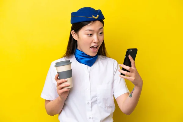 Airplane Chinese woman stewardess isolated on yellow background holding coffee to take away and a mobile