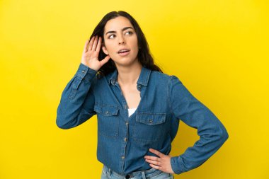Young caucasian woman isolated on yellow background listening to something by putting hand on the ear clipart