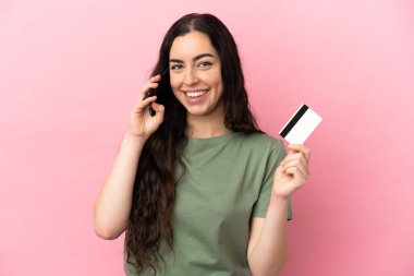 Pembe arka planda izole edilmiş genç beyaz bir kadın cep telefonuyla sohbet ediyor ve elinde bir kredi kartı tutuyor.