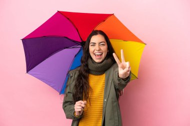 Young woman holding an umbrella isolated on pink background smiling and showing victory sign