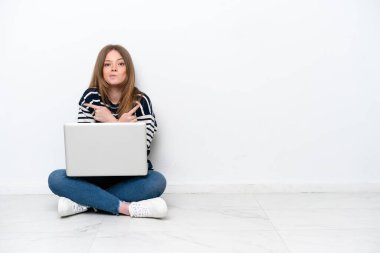 Young caucasian woman with a laptop sitting on the floor isolated on white background pointing to the laterals having doubts