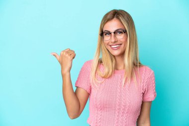 Sarışın Uruguaylı kız, mavi arka planda izole edilmiş bir şekilde bir ürün sunmak için kenarı işaret ediyor.