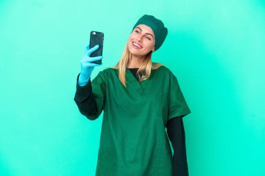 Young surgeon Uruguayan woman in green uniform isolated on blue background making a selfie