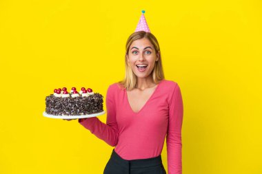 Blonde Uruguayan girl holding birthday cake isolated on yellow background with surprise facial expression