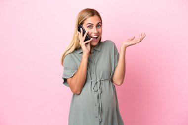 Genç Uruguaylı kadın mavi arka planda izole edilmiş hamile ve şaşırmış bir ifadeyle cep telefonu kullanıyor.