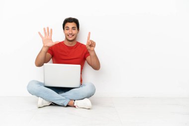 Caucasian handsome man with a laptop sitting on the floor counting six with fingers