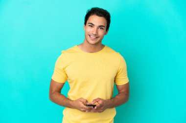 Kafkas yakışıklı adam izole mavi arka planda cep telefonuyla bir mesaj gönderiyor.