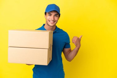Yalıtılmış sarı duvarın üstünde teslimatçı bir ürün sunmak için yan tarafı işaret ediyor.