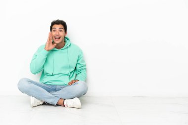 Caucasian handsome man sitting on the floor shouting with mouth wide open