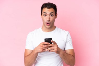 Young caucasian man isolated on pink background looking at the camera while using the mobile with surprised expression