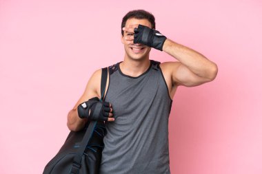 Young sport caucasian man with sport bag isolated on pink background covering eyes by hands and smiling
