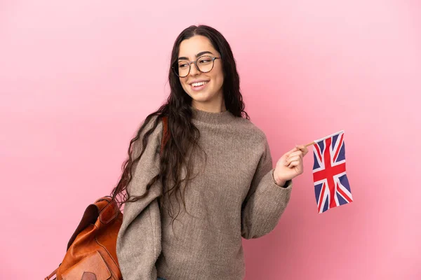 Young caucasian woman holding an United Kingdom flag isolated on pink background looking to the side and smiling